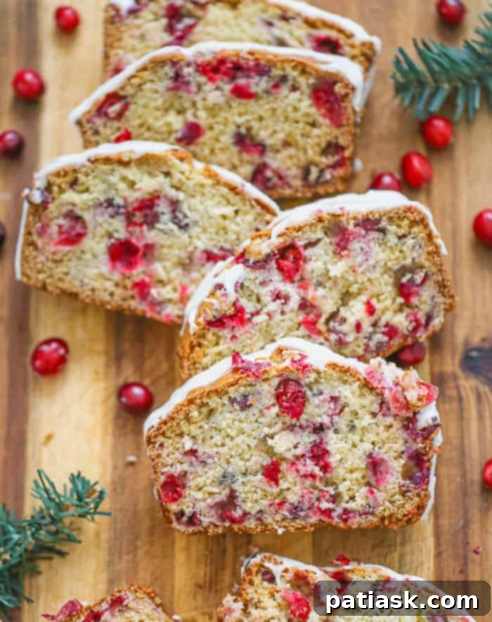 A sliced loaf of cranberry orange quick bread drizzled with an orange glaze, on a wooden cutting board.