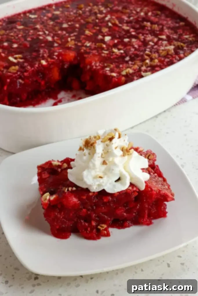 A festive cranberry jello salad, studded with fruit and nuts, in a clear glass bowl.